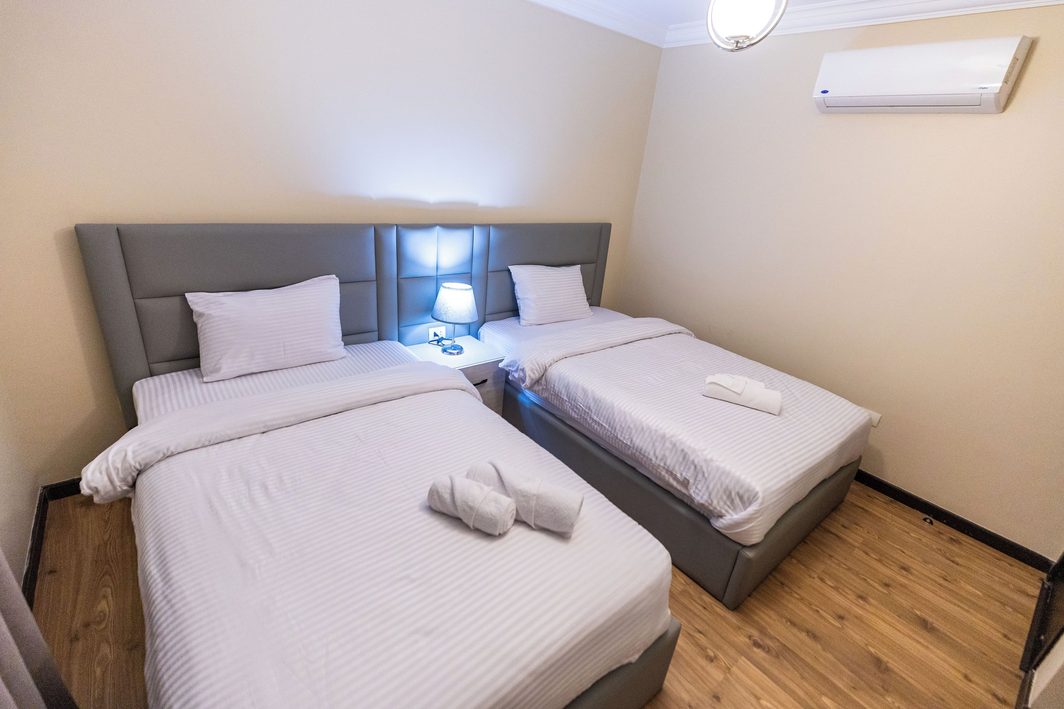 A hotel room with two beds, a nightstand with a lamp, and an air conditioning unit on the wall.