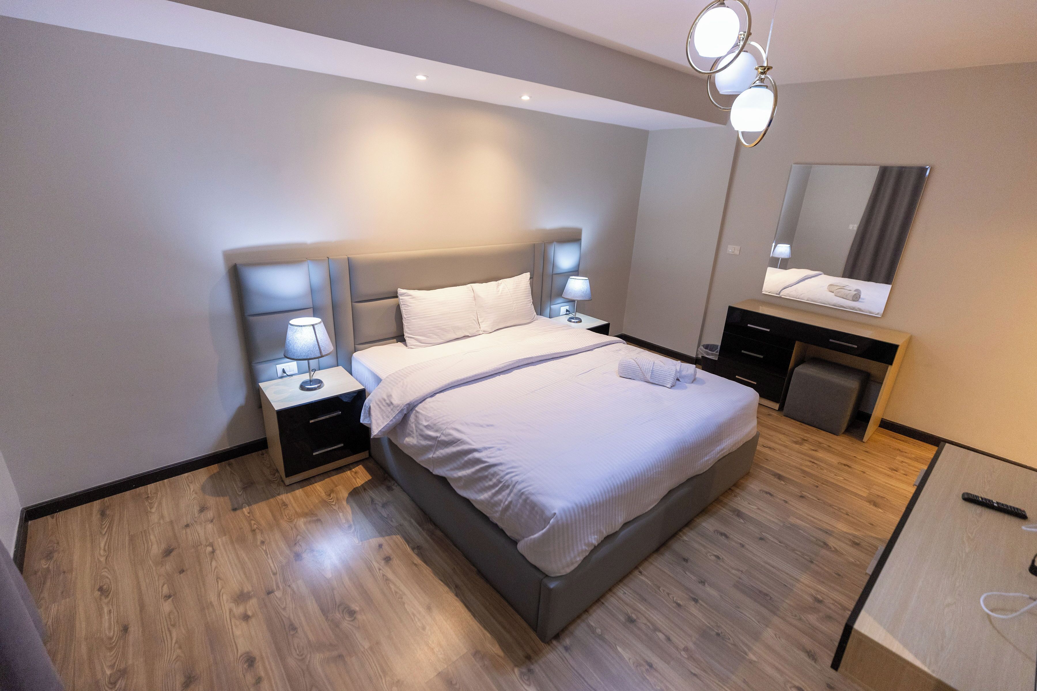 A modern bedroom with a large bed, bedside tables with lamps, a dresser, and a mirror on the wall.