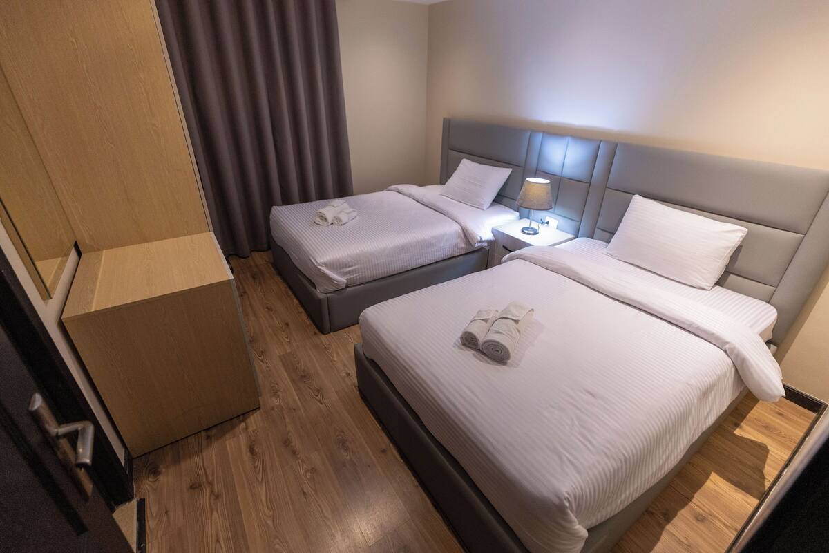 A hotel room with two beds, a nightstand, a lamp, and a wooden floor.