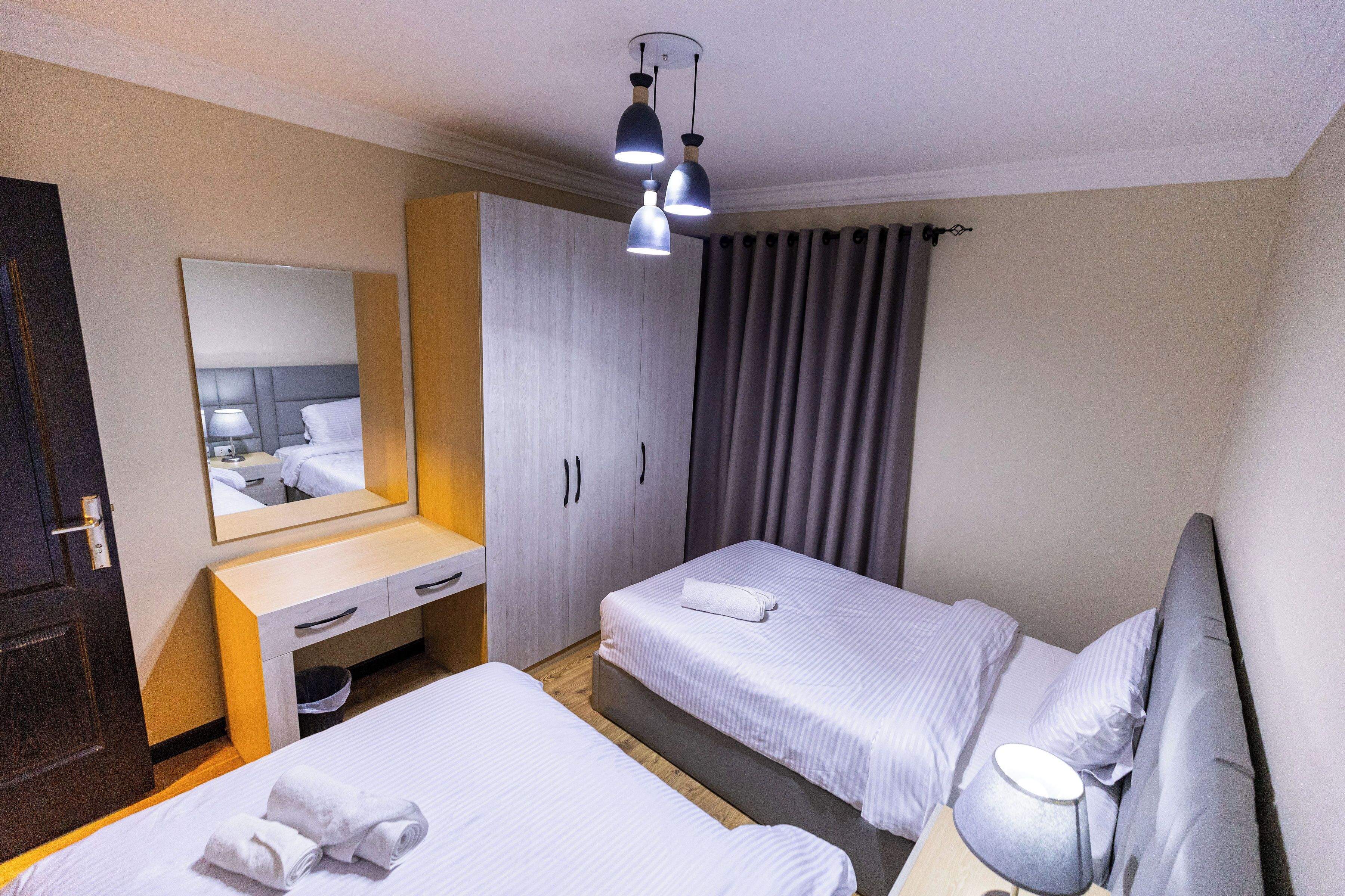 A hotel room with two beds, a wooden desk, a mirror, a wardrobe, and a window with curtains.