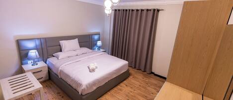 A modern bedroom with a large bed, bedside tables, a nightstand, a wooden floor, and a ceiling light fixture.