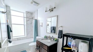 Un baño moderno con bañera, inodoro, lavabo y ducha. Cuenta con una ventana que proporciona luz natural, un perchero y un estante con artículos de aseo.
