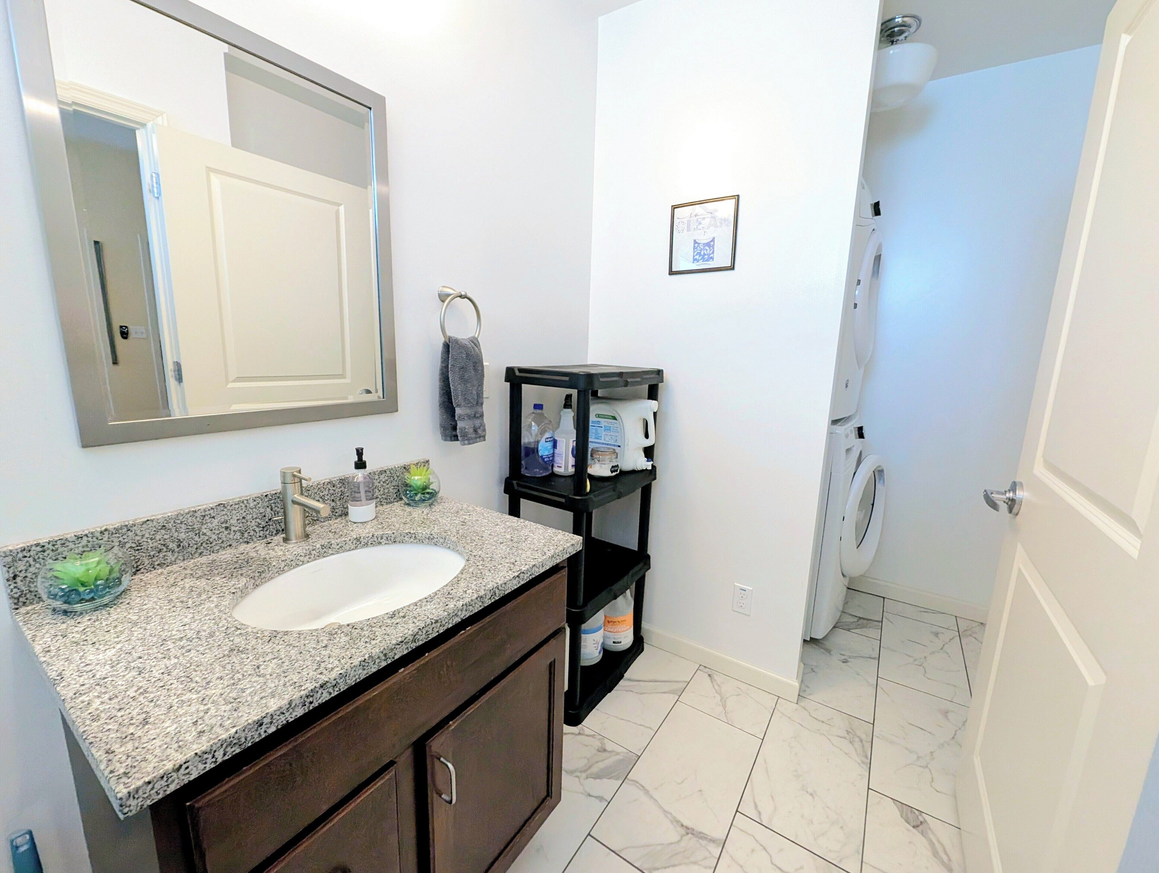 A bathroom with a granite countertop sink, a mirror, a towel rack, a small shelf with toiletries, and a shower area.