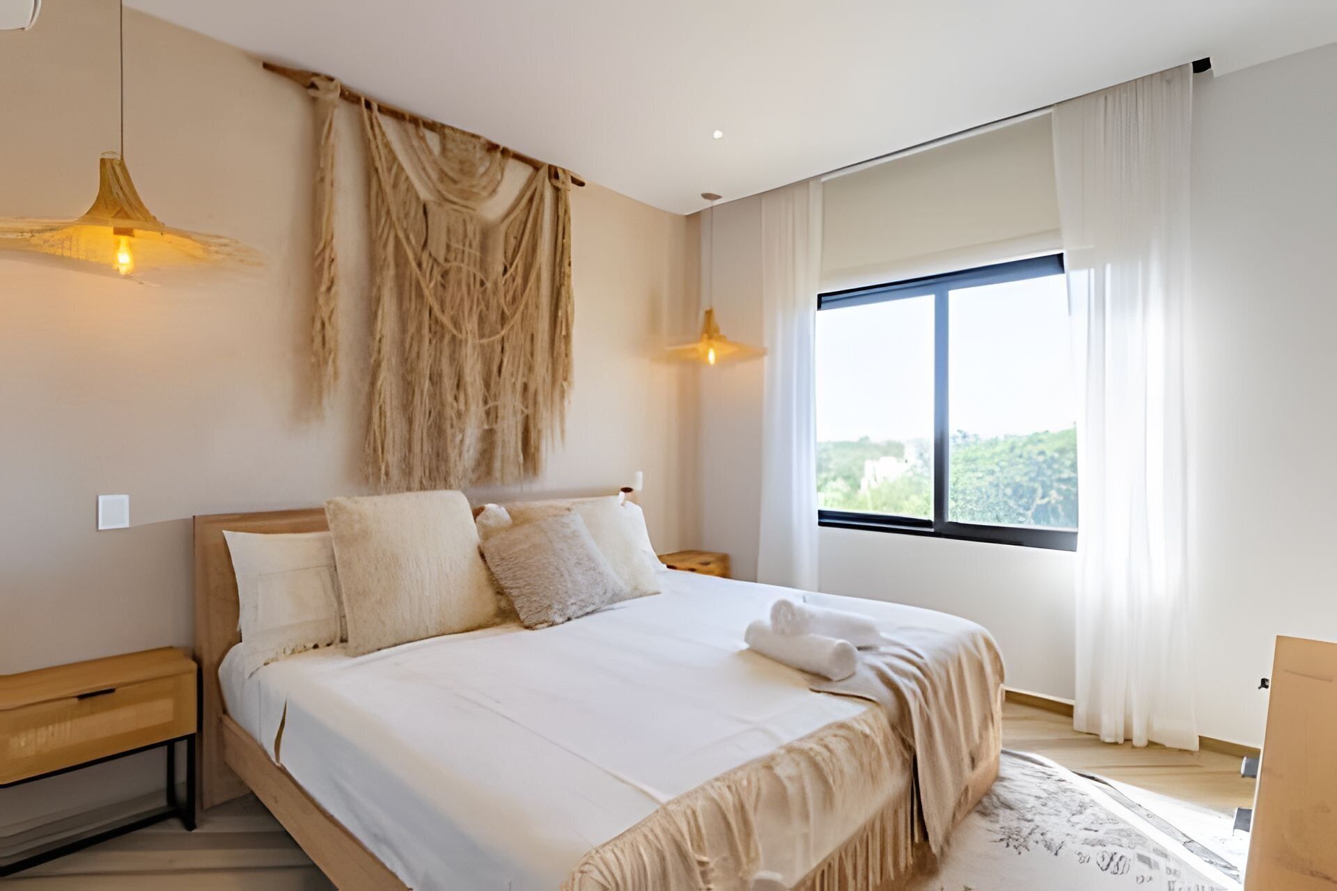 A modern bedroom with a large bed, two bedside lamps, a view of greenery outside, and a large window with curtains.
