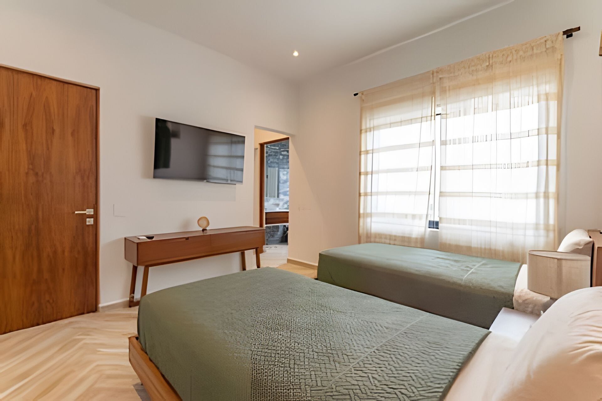 A bedroom with two beds, a wooden desk, a television, and a window with sheer curtains.
