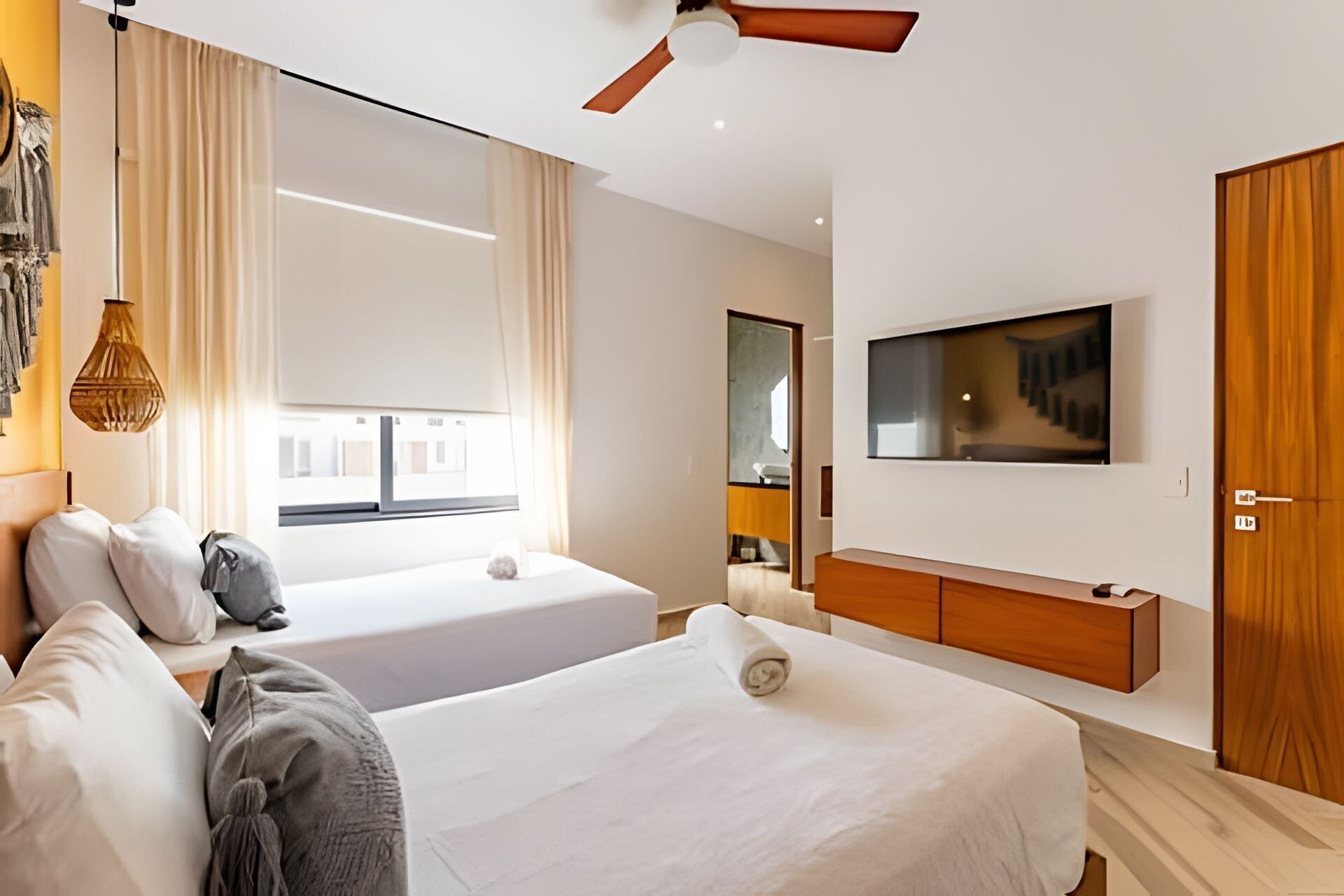 A modern hotel room with two beds, a large window, a ceiling fan, a TV, and a wooden wardrobe.