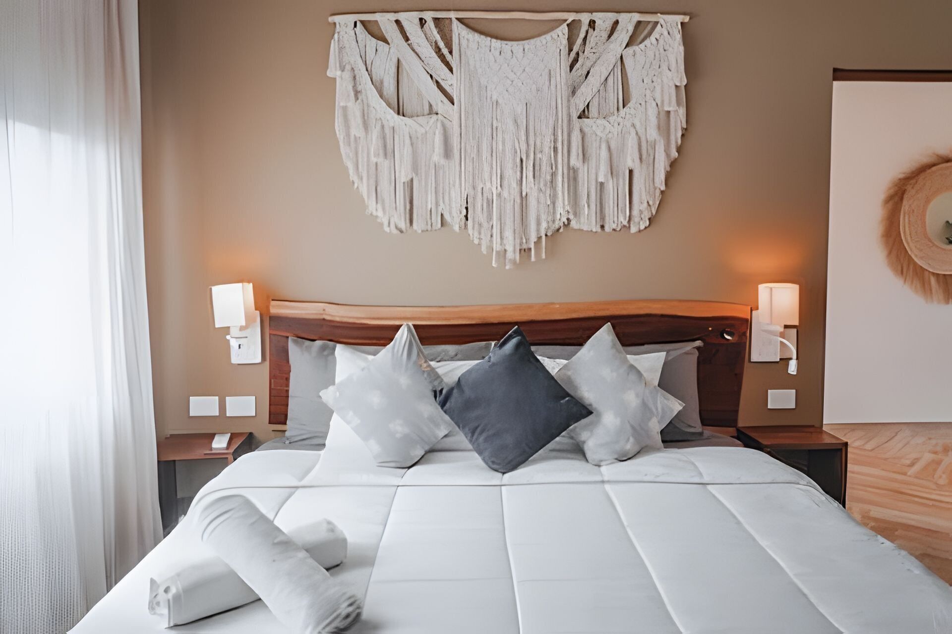 A neatly made bed with a white duvet and several pillows, a wooden headboard, bedside lamps, and a wall-mounted macrame piece.