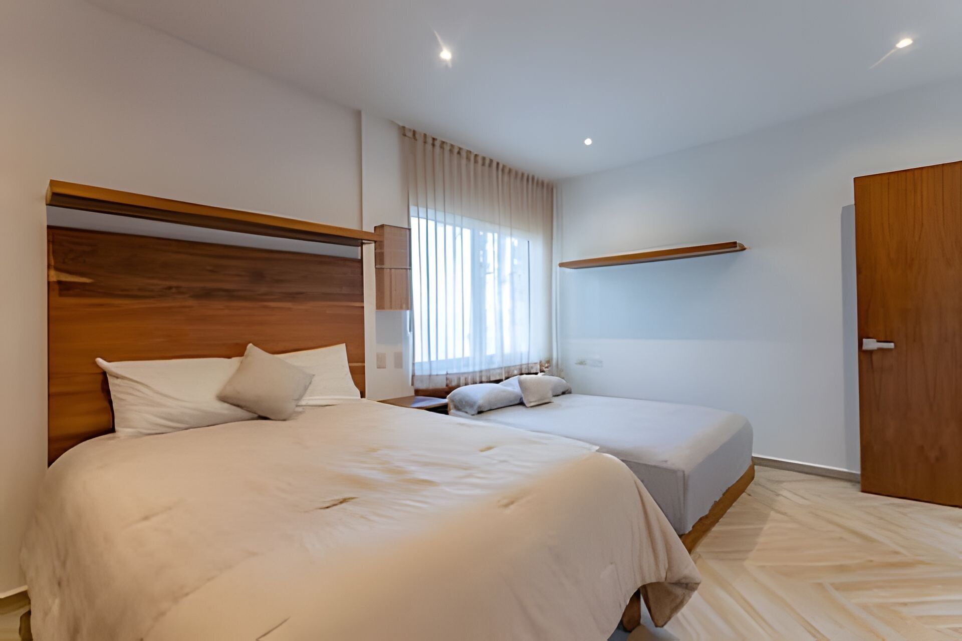 A modern hotel room with two beds, a wooden headboard, a large window with curtains, a wooden door, and a built-in shelf above the bed.