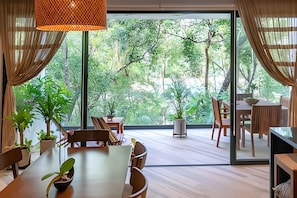 A spacious room with a large sliding glass door leading to a lush outdoor area. Inside, there is a dining table with chairs, a small table with a potted plant, and a wooden cabinet. - Oasis Yaaxlum 2BR (Tulum)