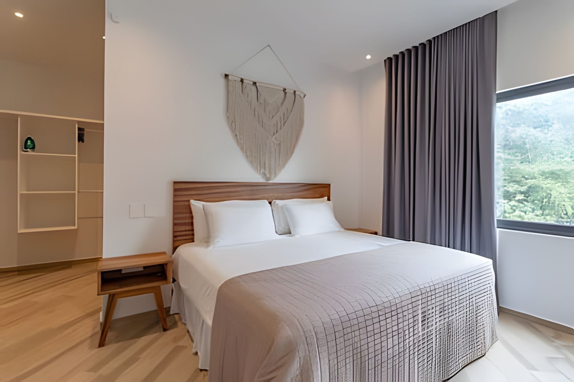 A modern bedroom with a wooden headboard, a large bed with white bedding, a bedside table, a hanging light fixture, and a view of greenery through a window with grey curtains.