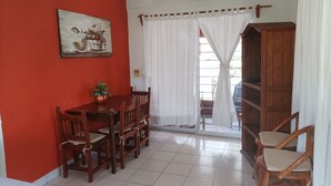 Un coin repas avec un mur rouge, une table en bois et des chaises. On y trouve également un ventilateur de plafond, une plante en pot et un rideau blanc.