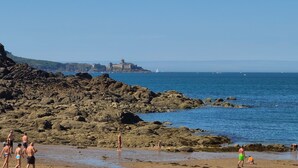 Ein felsiger Strand mit klarem, blauem Wasser, ein Schloss in der Ferne und Personen, die spazieren gehen.