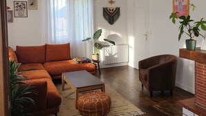 Ein Wohnzimmer mit einer Couch, einem Sessel, einem Hocker und einem Couchtisch. Es gibt Pflanzen, eine Pendelleuchte und Kunstwerke an den Wänden.