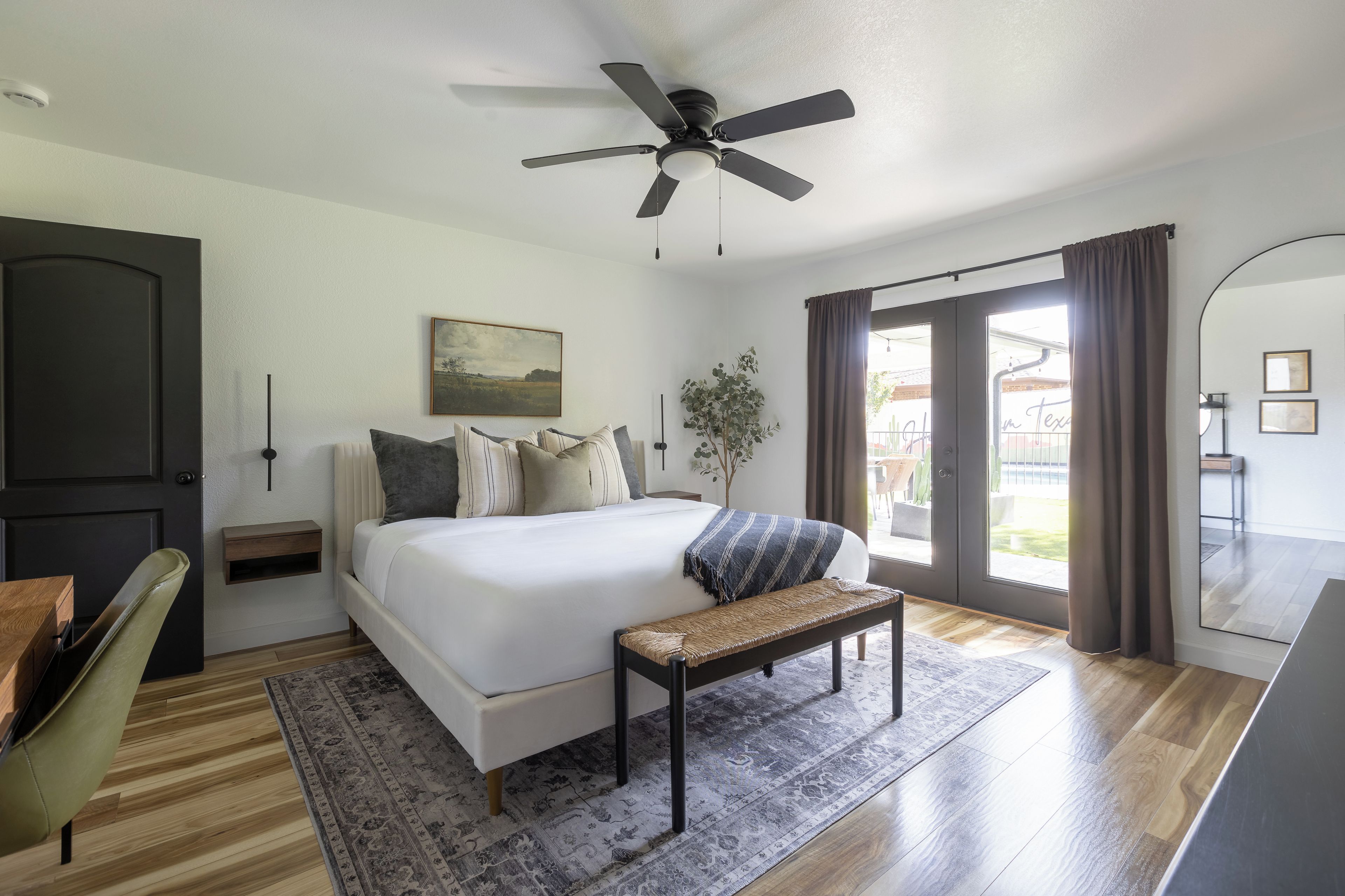 Un dormitorio moderno con una cama grande, un ventilador de techo, un banco, una mesita de noche, una silla y vistas al exterior.