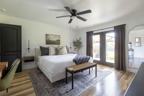 Une chambre moderne avec un grand lit, un ventilateur de plafond, un banc, une table de chevet, une chaise et une vue sur l’extérieur.