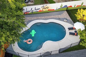 Ein Pool mit ungewöhnlicher Form, umgeben von einem weißen Zaun mit Wandmalerei, ausgestattet mit Liegestühlen und Sonnenschirm.