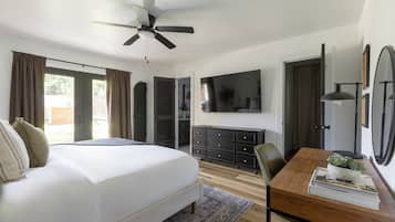 Une chambre avec un grand lit, un ventilateur de plafond, un téléviseur fixé au mur, une commode en bois et un bureau agrémenté d’une plante.
