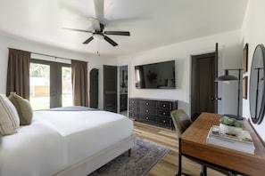 A bedroom with a large bed, a ceiling fan, a TV mounted on the wall, a wooden dresser, and a desk with a plant.