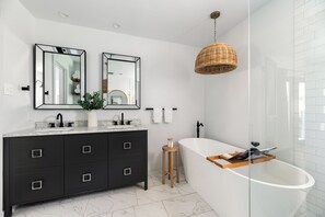 Ein modernes Badezimmer mit dunklem Waschtisch, einer freistehenden Badewanne und einer Korbstuhl-Pendelleuchte.