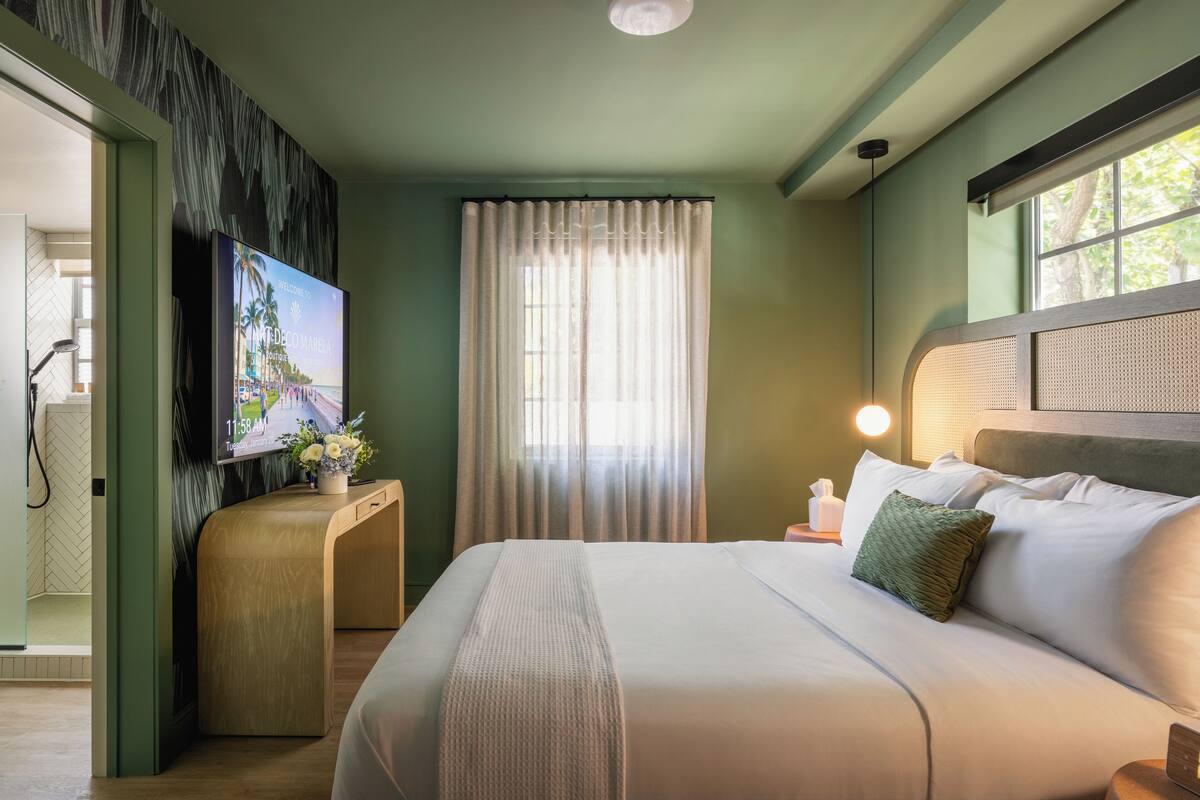 A hotel room with a bed, a TV, a window with curtains, and a bedside table with a lamp.
