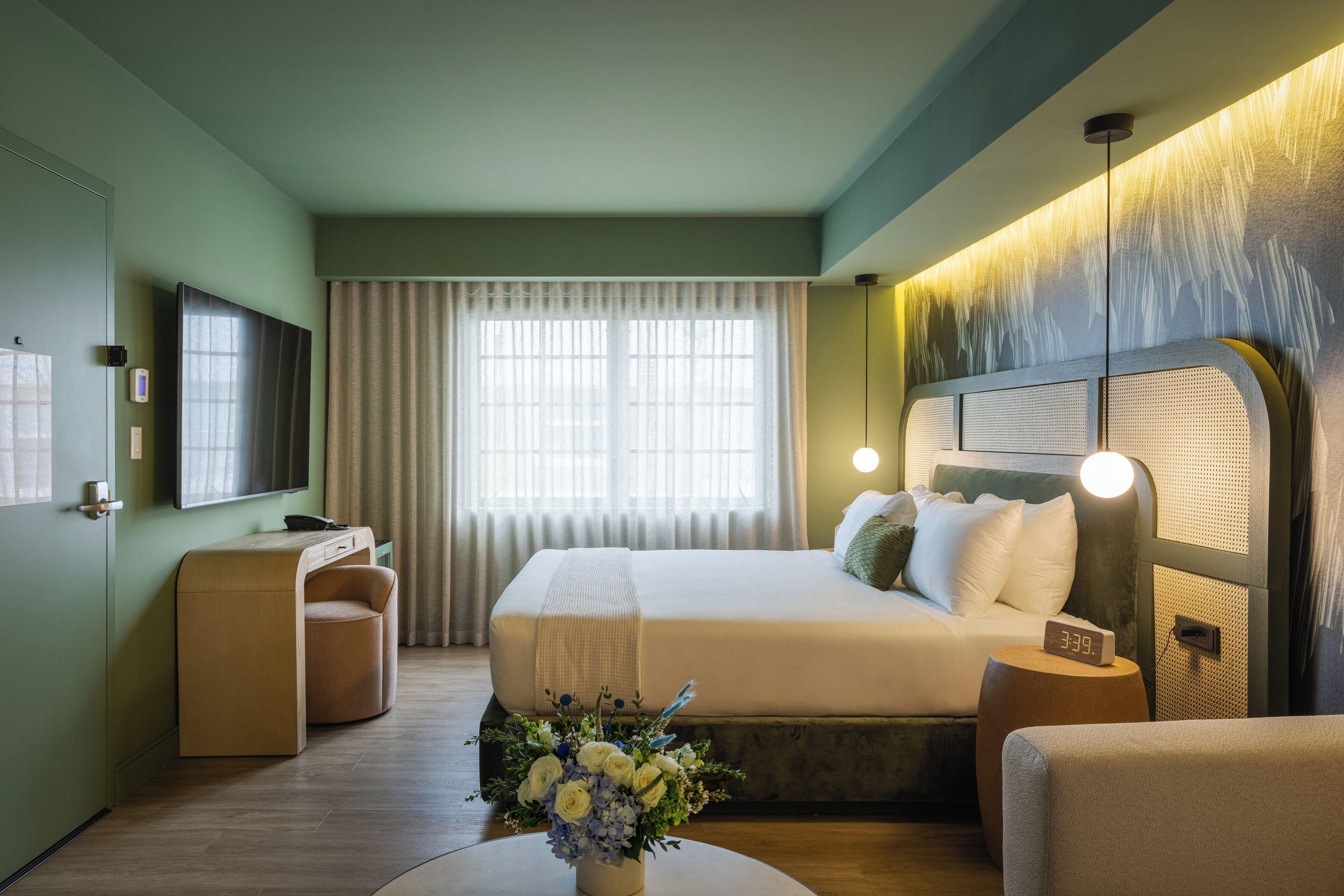 A hotel room with a bed, a desk, a chair, a bedside table, a vase of flowers, and a wall-mounted TV.