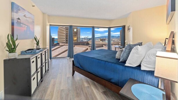 A modern hotel room with a large bed, a desk, and a view of the city through floor-to-ceiling windows.