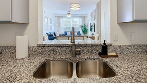 Une cuisine équipée d’un évier à deux bacs, d’un plan de travail en granit et offrant une vue sur un espace de vie comprenant un canapé et un ventilateur de plafond.