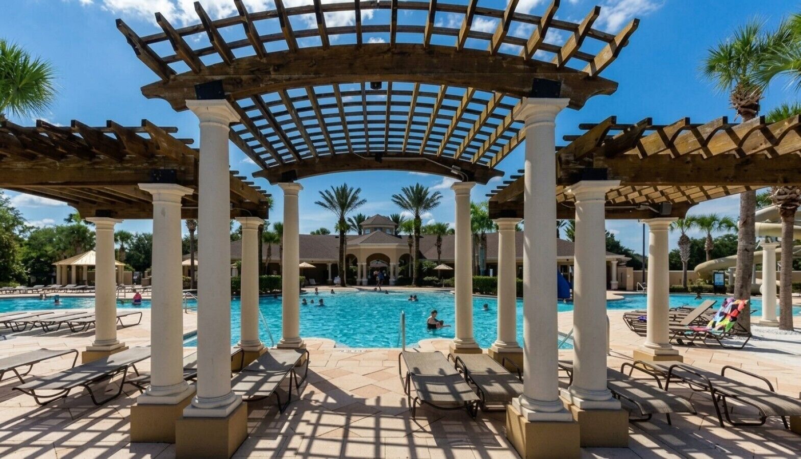 Área de piscina com estrutura de pérgola, cadeiras de descanso e piscina azul cristalina.