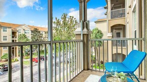 Un balcon avec une chaise bleue, une petite table avec un plateau, et une vue sur un parking et des immeubles.