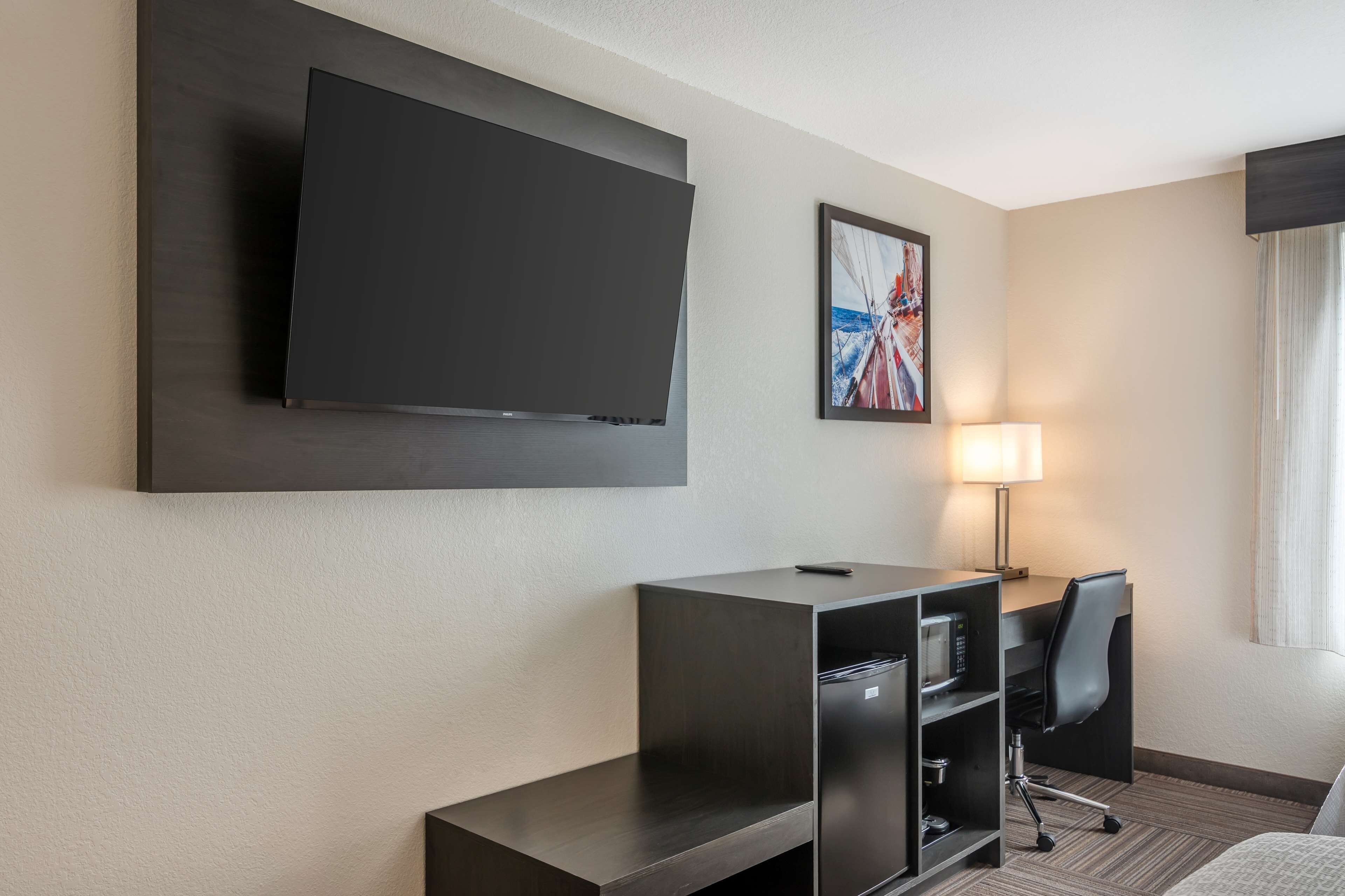 A hotel room with a large flat-screen TV mounted on the wall, a desk with a chair, a lamp, and a bed.