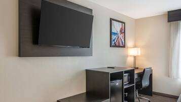 A hotel room with a large flat-screen TV mounted on the wall, a desk with a chair, a lamp, and a bed.