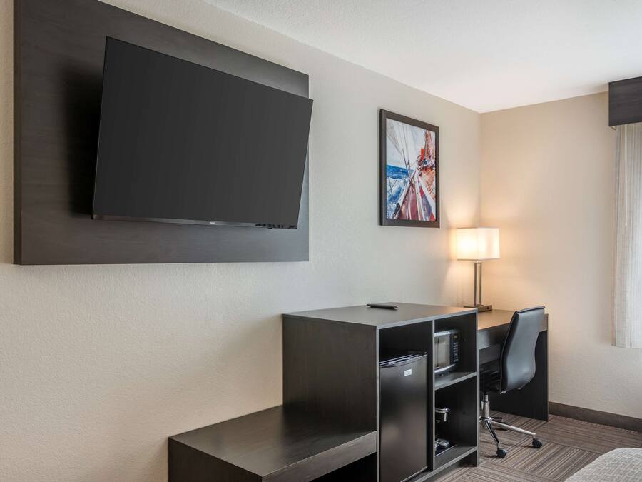 A hotel room with a large flat-screen TV mounted on the wall, a desk with a chair, a lamp, and a bed.