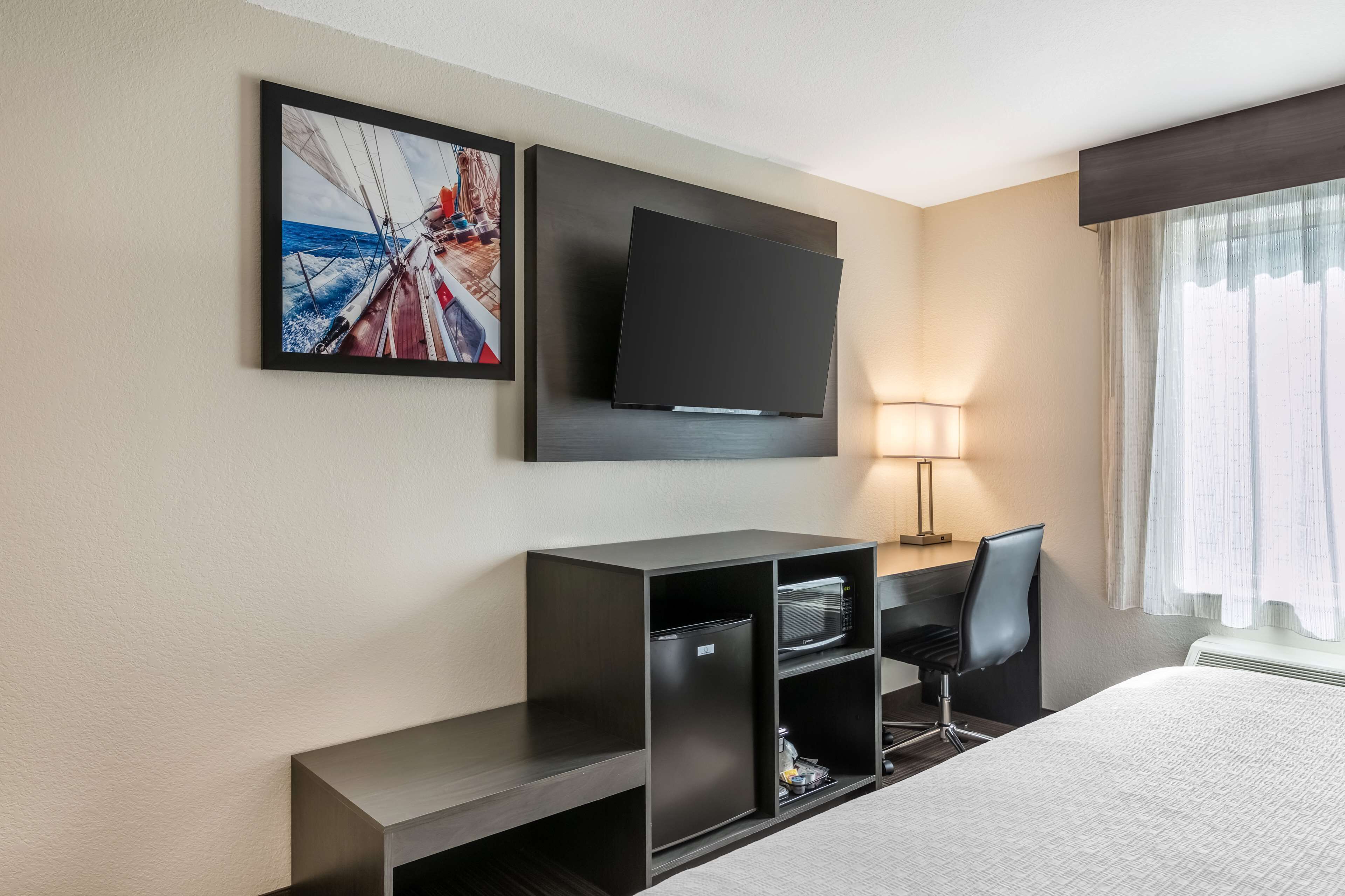 Une chambre d’hôtel équipée d’un lit, d’un téléviseur à écran plat fixé au mur, d’un bureau avec une chaise, d’un petit réfrigérateur et d’une fenêtre avec des rideaux.