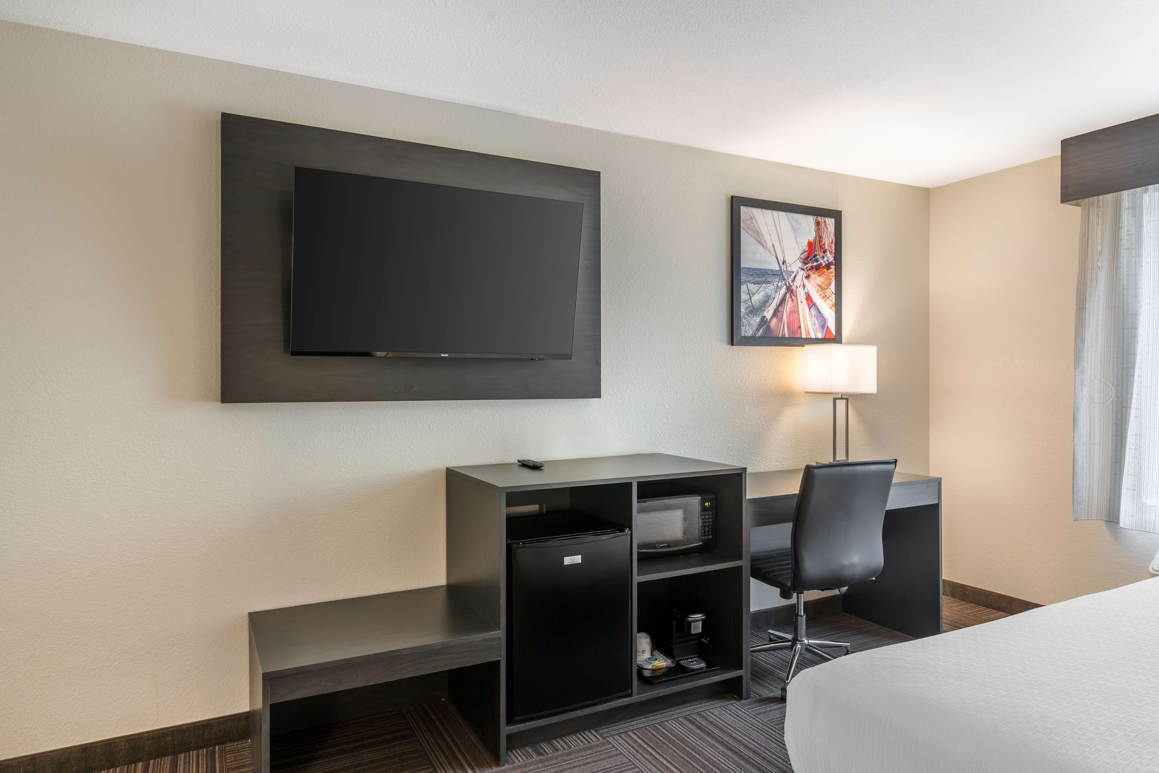 Quarto de hotel com televisão de ecrã plano na parede, uma secretária com cadeira, uma cama e um pequeno espaço de estar com uma lâmpada.
