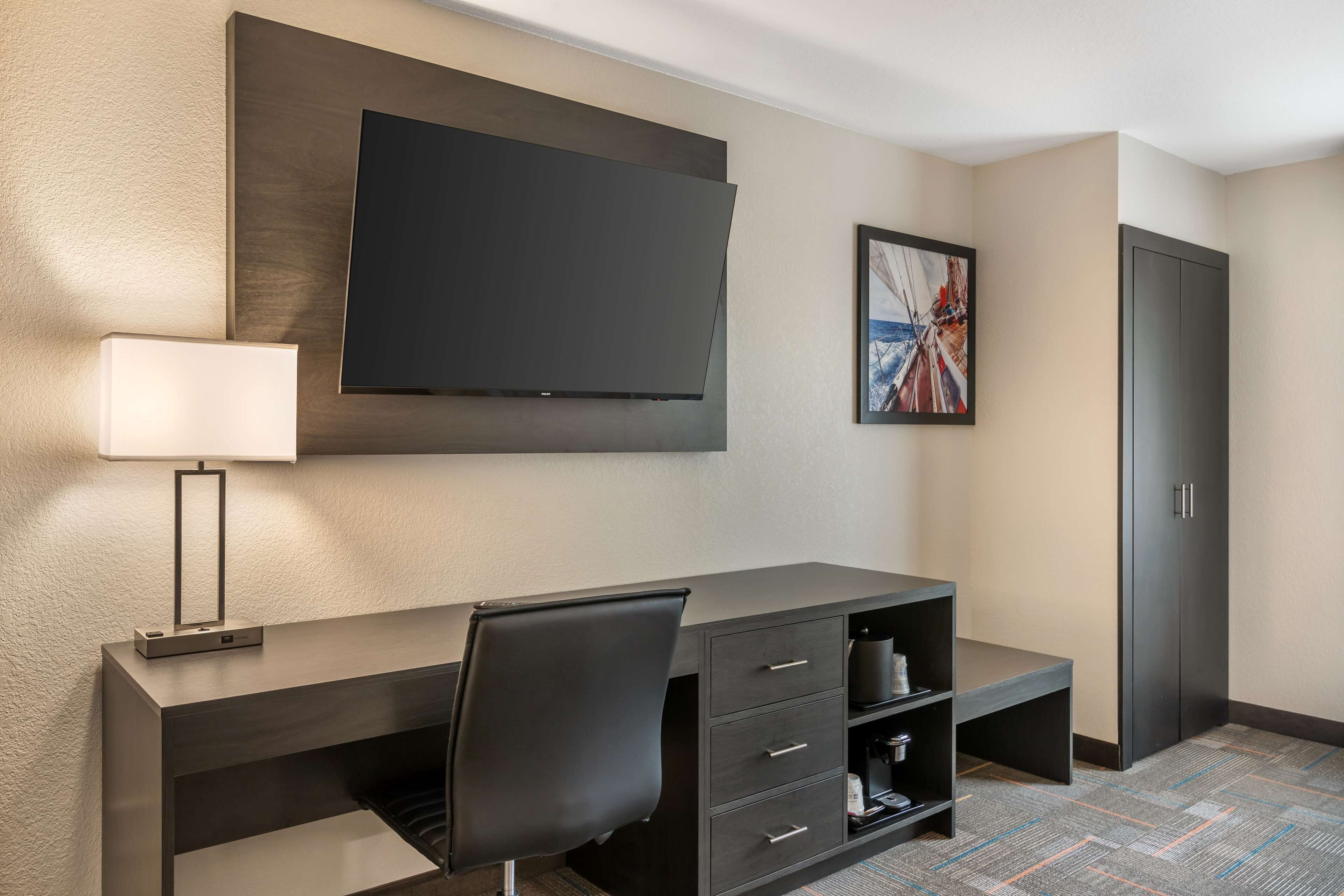 A modern hotel room with a large flat-screen TV mounted on the wall, a desk with a chair, a lamp, and a framed picture.