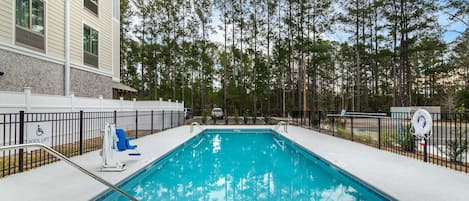 A rectangular swimming pool with a pool lift and a lifeguard chair, surrounded by a fence and trees.