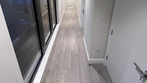 Een smalle gang met een lichtkleurige houten vloer, een glazen deur links en een witte deur rechts.