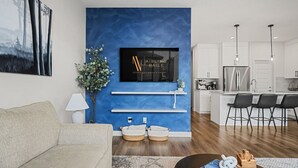 A modern living room with a blue accent wall, a flat-screen TV, a sofa, and a coffee table with books and a toy. - Wanderer`s Haven | Private Nordic Spa | Game Room (Cochrane)