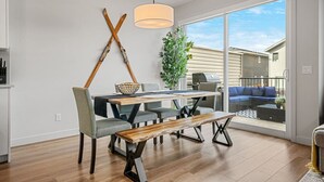 A modern dining area with a wooden table, grey upholstered chairs, and a bench. There is a large window with a view of neighboring houses. - Wanderer`s Haven | Private Nordic Spa | Game Room (Cochrane)