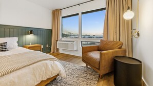A hotel room with a bed, a leather armchair, a standing lamp, a large window with curtains, and an air conditioning unit. - Comfort, Style & Views | Steps to Tours & Eats (Juneau)