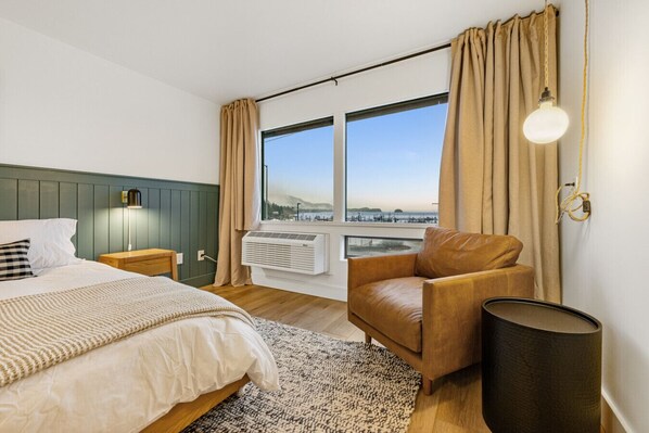 Une chambre d’hôtel comprenant un lit, un fauteuil en cuir, une lampe sur pied, une grande fenêtre avec des rideaux et un appareil de climatisation.