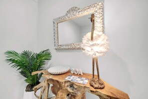 Una consola de madera con un espejo decorativo, un cuenco blanco con forma de concha y una lámpara con pantalla de plumas.