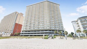 Un hotel frente a la playa con varios pisos, balcones y una playa arenosa en primer plano.