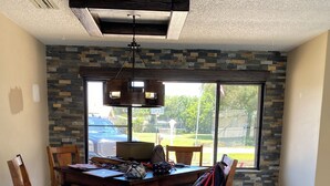 A dining area with a stone accent wall, a large window, and a ceiling-mounted light fixture. - Rustic Elegance- Private Heated Salt pool, 2 bed, 2 bath, Fenced & Pet friendly (Port St Lucie)