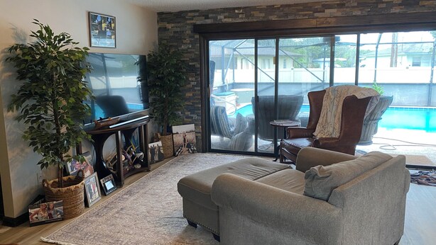 A living room with a grey sofa, a large potted plant, a flat-screen TV, a fireplace, and a view of a swimming pool. - Rustic Elegance- Private Heated Salt pool, 2 bed, 2 bath, Fenced & Pet friendly (Port St Lucie)