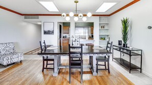 A dining area with a wooden table, chairs, and a modern chandelier. There is a sofa and a console table with a plant. The room has wooden flooring and white walls.