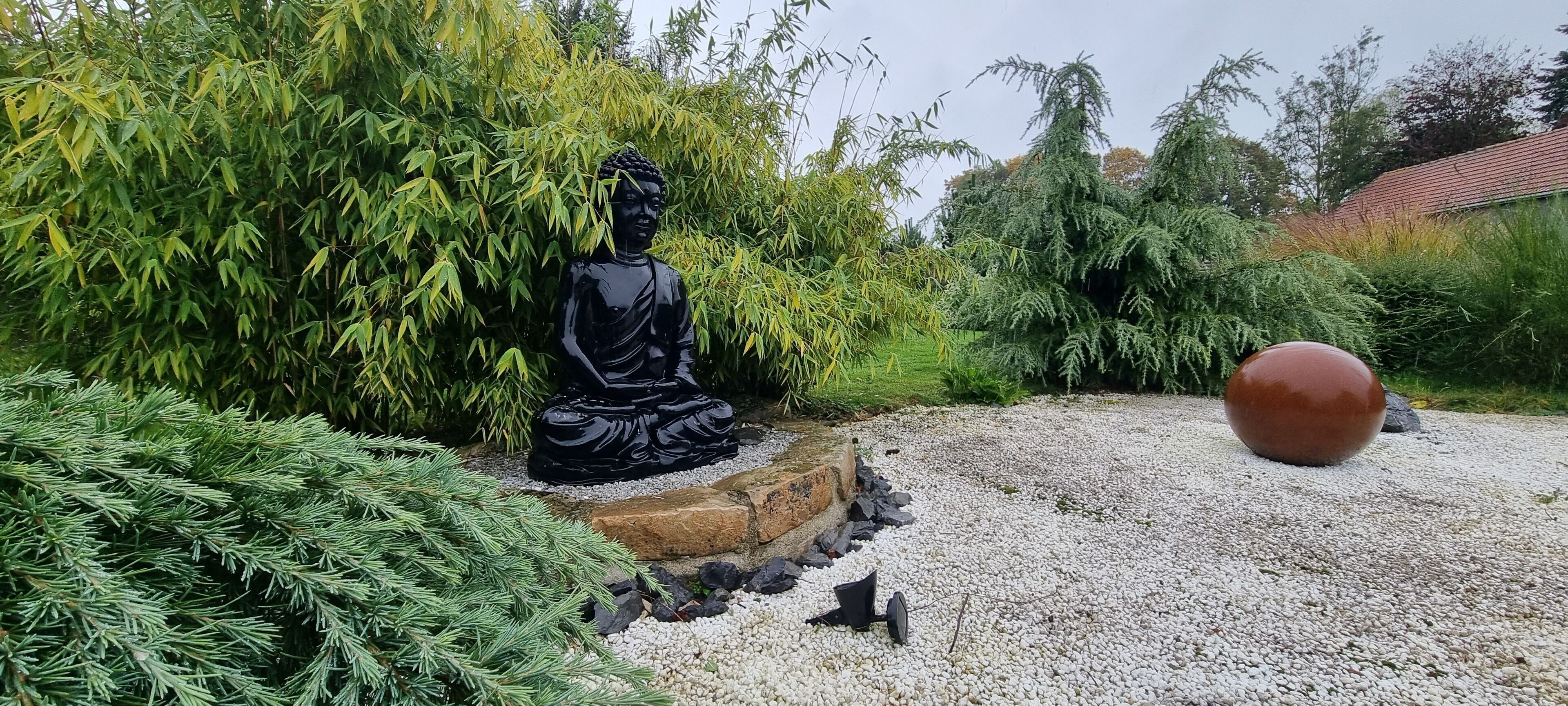 Una estatua negra de una figura sentada en un jardín con senderos de grava y follaje.