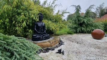 Una estatua negra de una figura sentada en un jardín con senderos de grava y follaje.