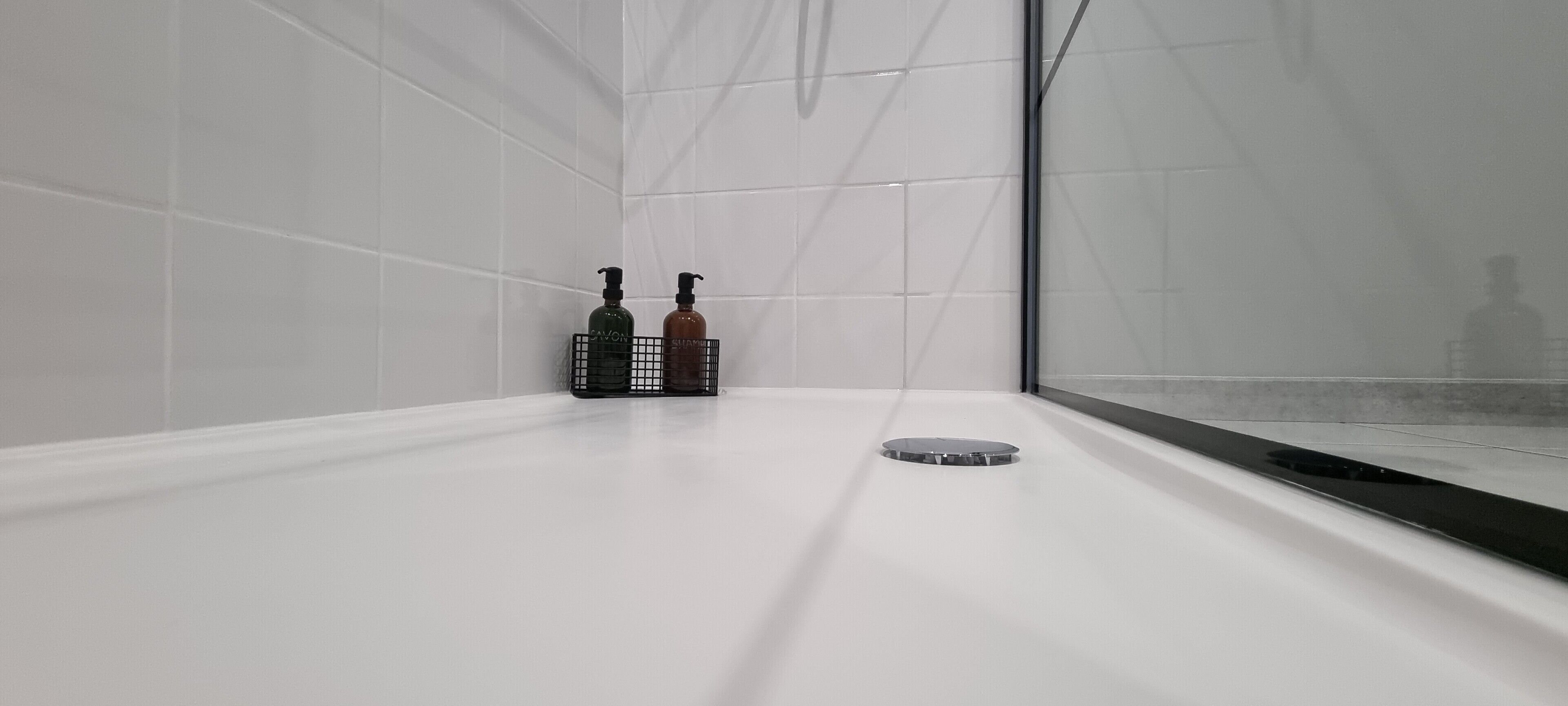 Une salle de bain moderne avec des carreaux blancs, une douche en verre et deux bouteilles de savon dans un panier noir.
