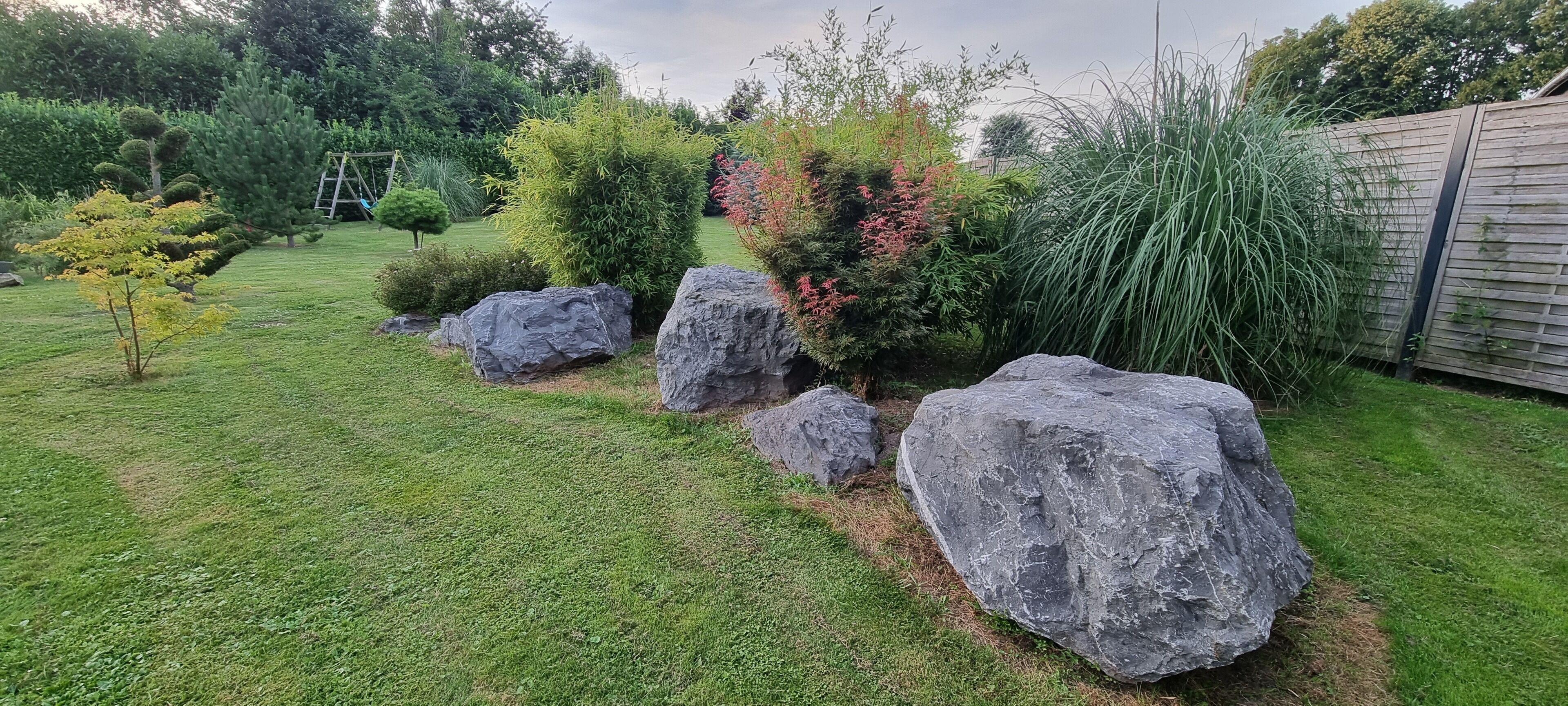 A well-maintained garden with large rocks, various shrubs, and a wooden fence.